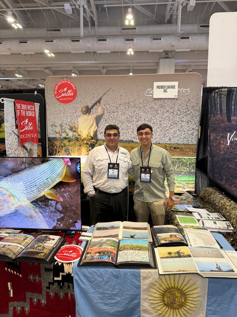 Two people beside photographs of hunting and fishing at a fair booth.